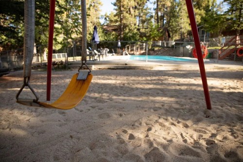 A yellow swing hangs in a sandy playground, with a pool and trees in the background.