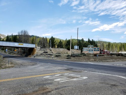 A rural intersection with a railway bridge overhead, surrounded by trees and signs for local businesses.