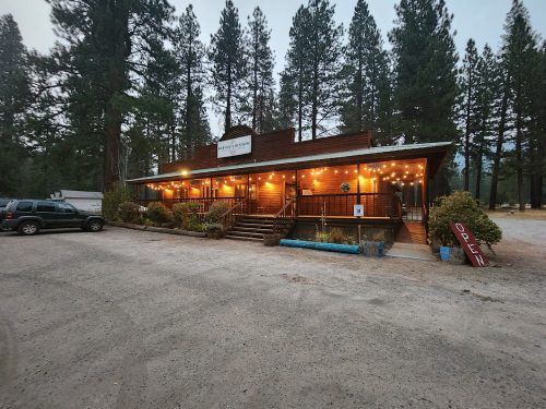 A rustic building with warm lights, surrounded by tall trees, featuring an "Open" sign and a gravel parking area.