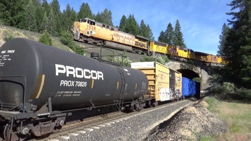 A Union Pacific train travels through a forested area, with freight cars and a tank car visible in the foreground.