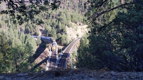 A train curves around a mountainous landscape, surrounded by dense green trees and rocky terrain.