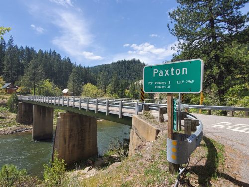 A green sign for Paxton near a bridge over a river, surrounded by trees and mountains under a clear blue sky.