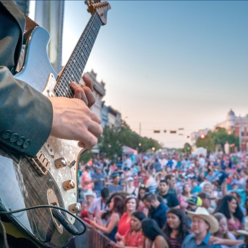 A guitarist performs on stage in front of a large, enthusiastic crowd during an outdoor event at sunset.
