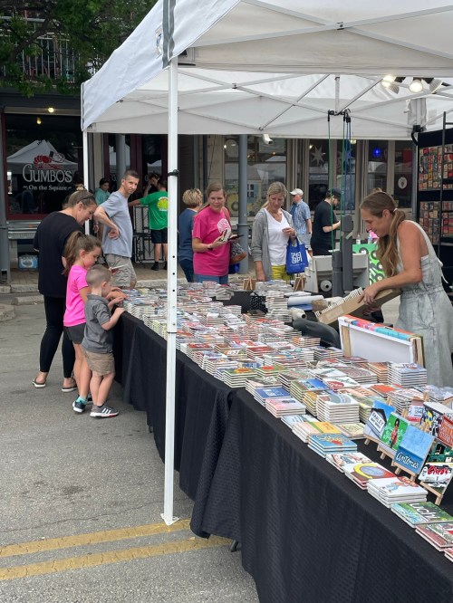 A busy outdoor market with people browsing colorful merchandise at vendor tables under white tents.