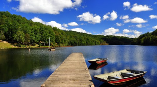A serene lake scene with two boats by a wooden dock, surrounded by lush green trees and a blue sky with fluffy clouds.