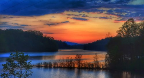 A serene sunset over a calm lake, with vibrant orange and blue hues reflecting on the water and silhouetted trees.