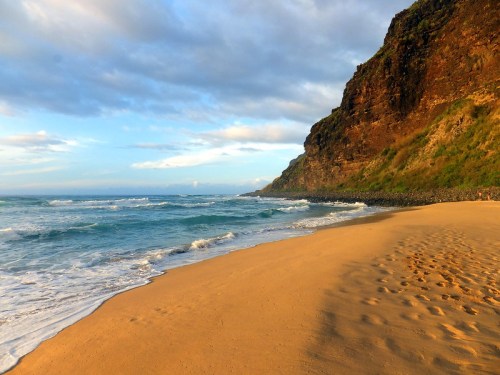 A serene beach scene with golden sand, gentle waves, and a rocky cliff under a partly cloudy sky.