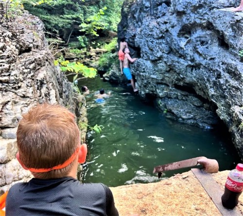 A child watches as others climb a rocky area near a swimming hole surrounded by trees.