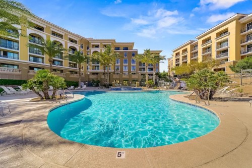 A sunny pool area surrounded by palm trees and buildings, featuring lounge chairs and a clear blue sky.