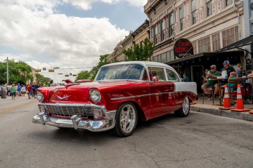 A classic red and white 1956 car parked on a street with people and buildings in the background.