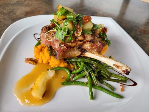 A plated dish featuring a grilled pork chop on sweet potato puree, topped with greens, apples, and green beans.