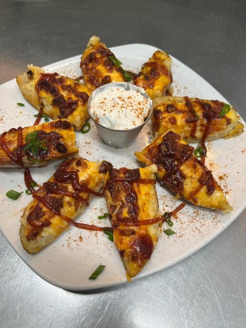 A plate of cheesy potato wedges topped with barbecue sauce and green onions, served with a side of ranch dressing.