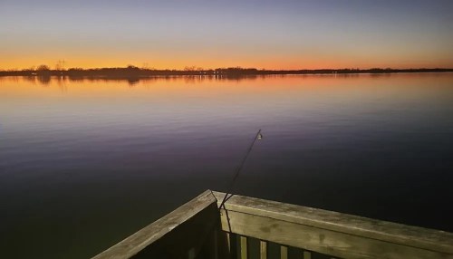 A serene lake at sunset, with calm waters reflecting vibrant colors and a fishing rod resting on a wooden dock.