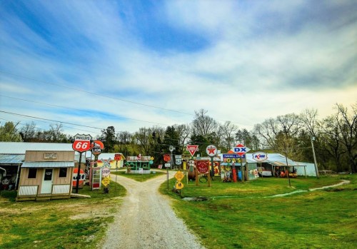 A scenic view of a gravel road lined with vintage gas station signs and buildings under a cloudy sky.