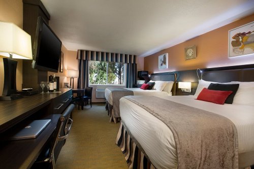 A cozy hotel room with two beds, a desk, a TV, and a window with natural light. Modern decor and warm colors.