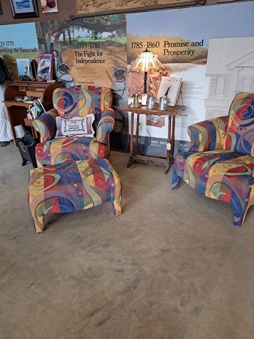 Two colorful patterned chairs with a small table and lamp between them, set against a wall with historical posters.