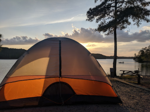 A tent by a lake at sunset, with a silhouette of a boat in the distance and trees framing the scene.