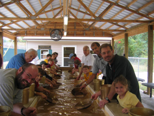 A group of adults and children panning for gold in a covered outdoor area, smiling and engaged in the activity.