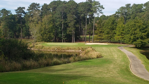 A scenic view of a golf course with a pond, sand traps, and surrounded by tall trees under a clear sky.