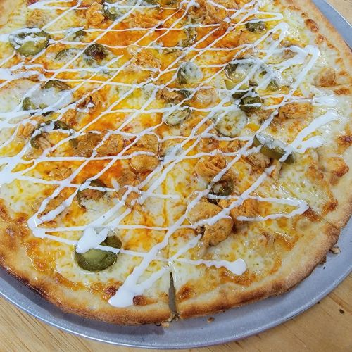 A close-up of a pizza topped with cheese, pickles, chicken, and drizzled with ranch dressing on a silver tray.