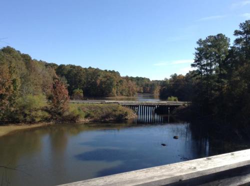 A serene river scene with a wooden bridge, surrounded by trees in autumn colors under a clear blue sky.