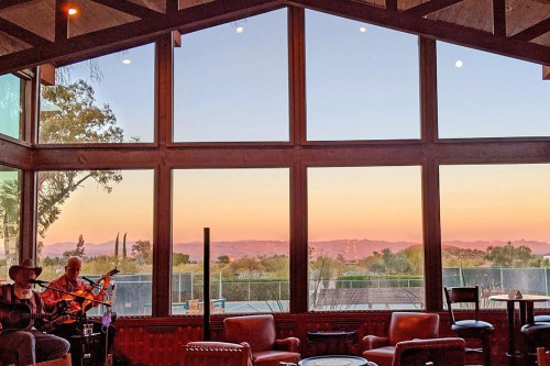 A cozy interior with musicians playing, framed by large windows showcasing a sunset over distant mountains.