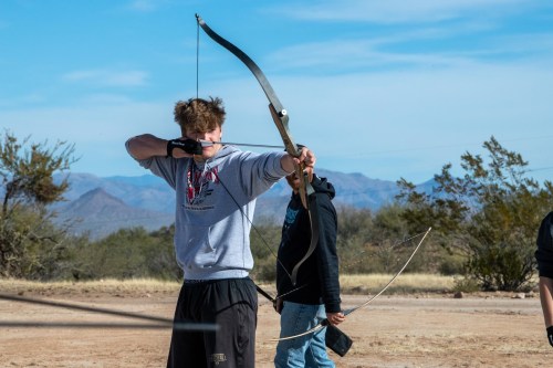 A young man aims a bow and arrow at a target, with mountains and trees in the background. Another person stands nearby.