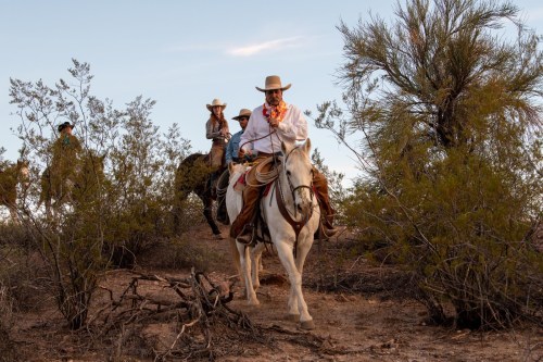 A group of horseback riders traverses a desert landscape, with one rider in a white shirt and hat leading the way.