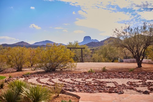 A desert landscape featuring a stone labyrinth, shrubs, and distant mountains under a partly cloudy sky.