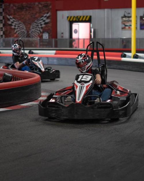 Two go-karts racing on a track, one with the number 19 and the other with the number 7, in a vibrant indoor setting.