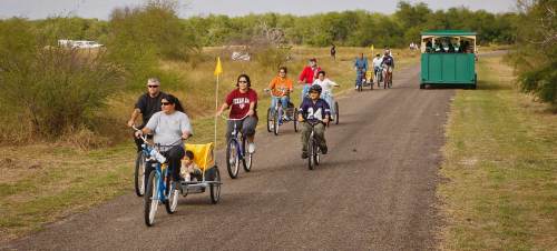 A group of people riding bikes on a path, with one bike pulling a small cart and a green vehicle in the background.