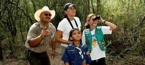 A group of four people, including children, observing nature in a wooded area, using binoculars and pointing.