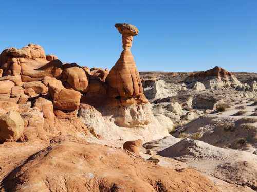 A unique rock formation resembling a hoodoo stands tall against a clear blue sky in a desert landscape.