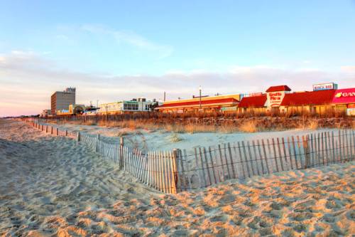 A sandy beach with a wooden fence, lined by buildings and restaurants under a colorful sunset sky.