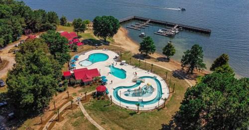 Aerial view of a pool area with water slides, surrounded by trees and a lake, with boats docked nearby.