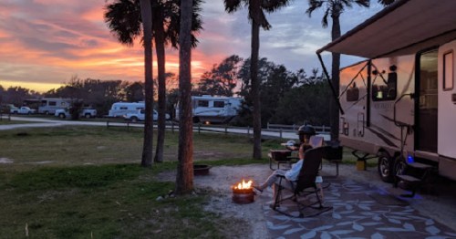 A person relaxes by a campfire outside an RV, with palm trees and a colorful sunset in the background.