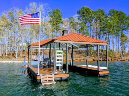 A wooden dock with a red roof, featuring an American flag, surrounded by water and trees under a clear blue sky.