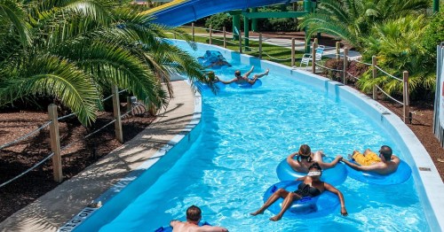 People enjoying a lazy river ride with colorful inner tubes surrounded by palm trees and a water slide in the background.