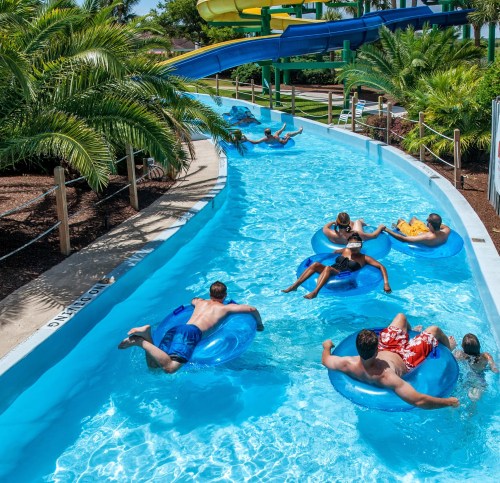 People floating on inner tubes in a lazy river at a water park, surrounded by palm trees and water slides.