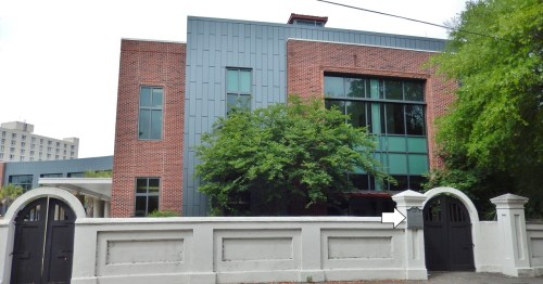 A modern building with a brick facade and large windows, surrounded by a white wall and greenery.