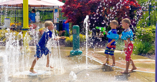 Three children play in a splash pad, laughing and spraying water with toys on a sunny day.