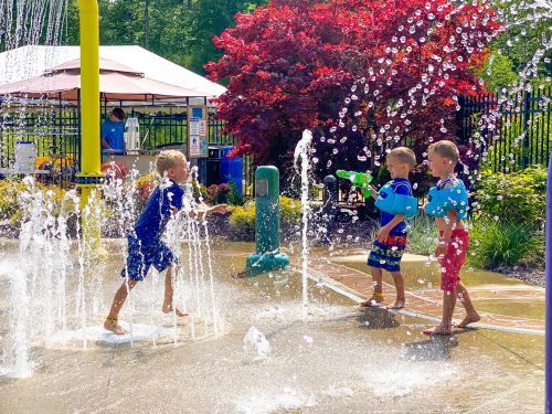 Three children play in a splash pad, spraying water and enjoying a sunny day surrounded by colorful flowers and trees.