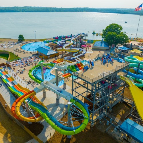 Aerial view of a water park featuring colorful slides, pools, and sunbathers by a lake on a sunny day.