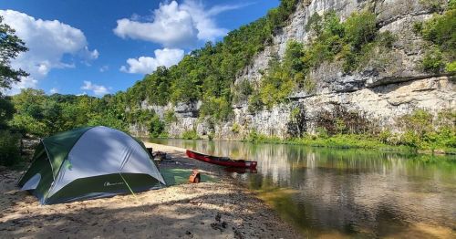 A tent by a calm river, surrounded by lush greenery and rocky cliffs under a blue sky with fluffy clouds.