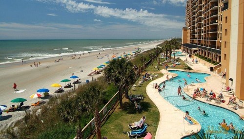 A sunny beach scene with people relaxing by the pool and on the sand, surrounded by umbrellas and palm trees.