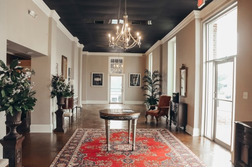 Elegant lobby with a chandelier, a round table, and decorative plants, featuring a red area rug and stylish furnishings.
