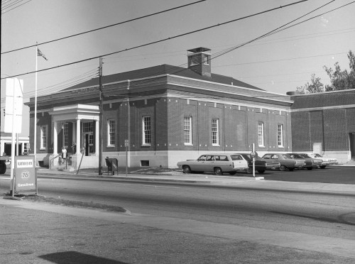 Historic brick building with a columned entrance, surrounded by parked cars and utility poles, on a sunny day.