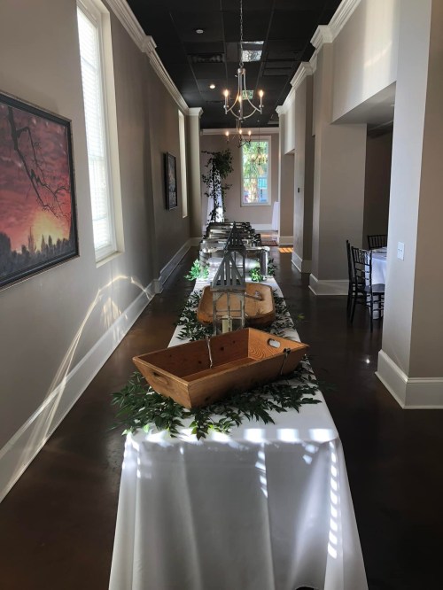 A long dining table decorated with wooden boats and greenery, set in a well-lit hallway with artwork on the walls.