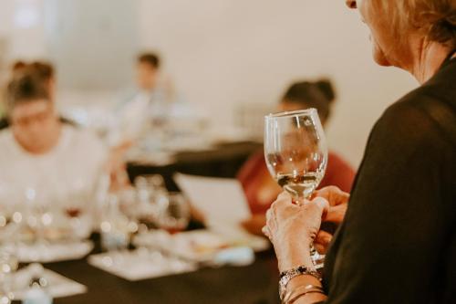 A person holding a wine glass in a blurred setting with others seated at a table, engaged in conversation.