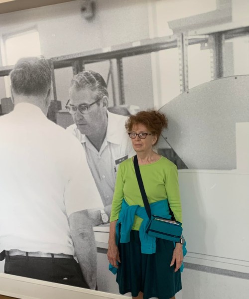 A woman in a green top and blue skirt stands in front of a black-and-white mural of two men at a counter.